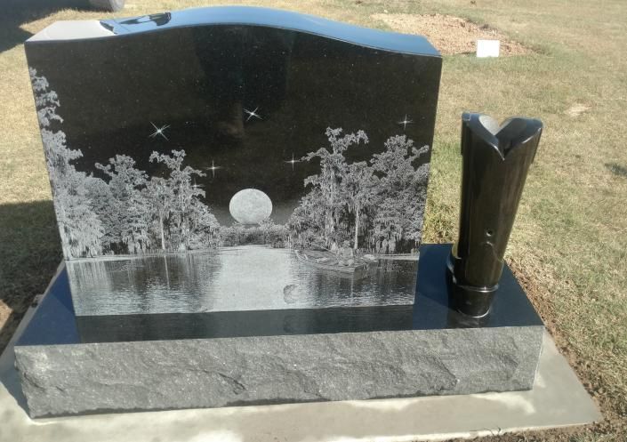 Black granite gravestone with etched moonlit trees and a vase on a concrete base in a cemetery