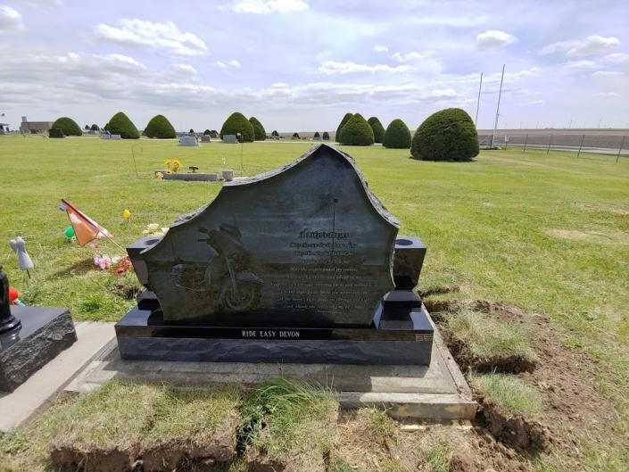Black memorial plaque on a concrete base in a grassy cemetery with trimmed shrubs and open fields.
