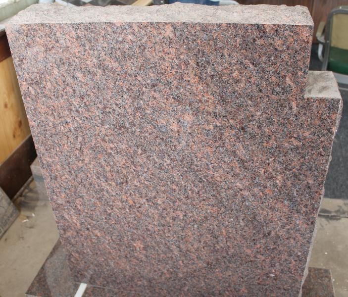 Red and gray speckled granite slab with a rough cut top edge, standing upright in a workshop.