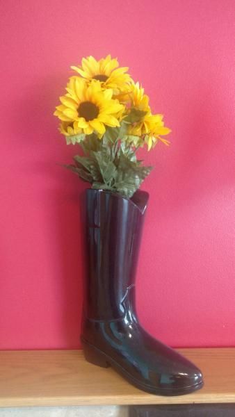 Yellow flowers arranged in a glossy black rain boot vase on a wooden shelf against a pink wall