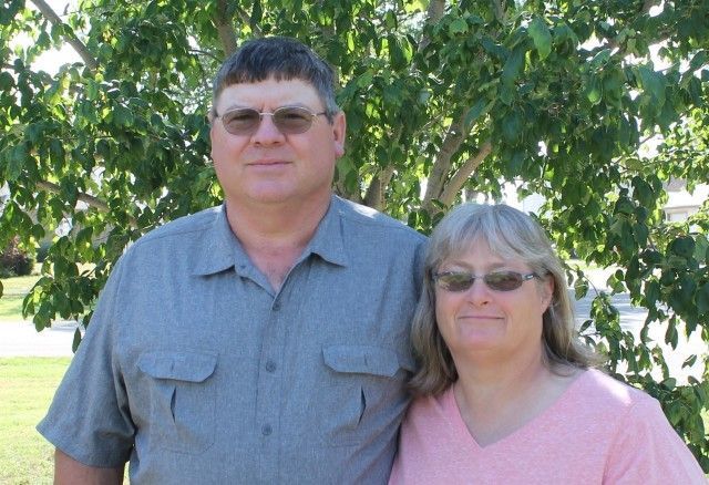 Two people standing outdoors under a leafy tree, wearing sunglasses and smiling at the camera.