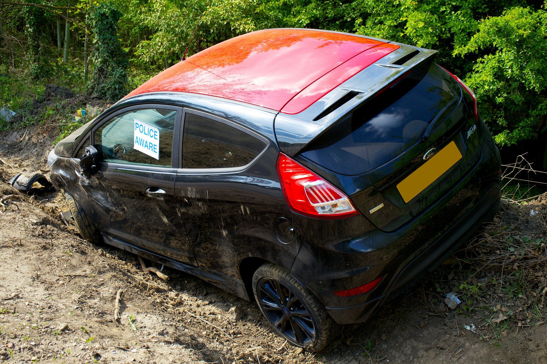 Black Ford Fiesta with red roof stuck in mud on the side of a road.