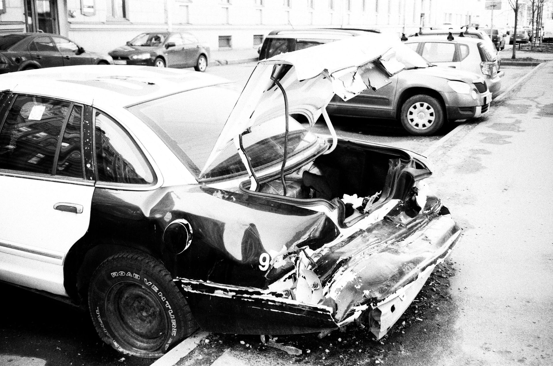 Damaged police car trunk, street scene. Rear-end collision visible, trunk open, other vehicles parked nearby.