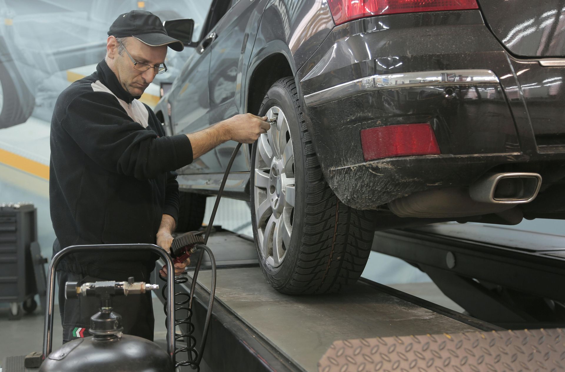 Mechanic inflating a tire on a black car in a garage. He wears a hat and glasses.