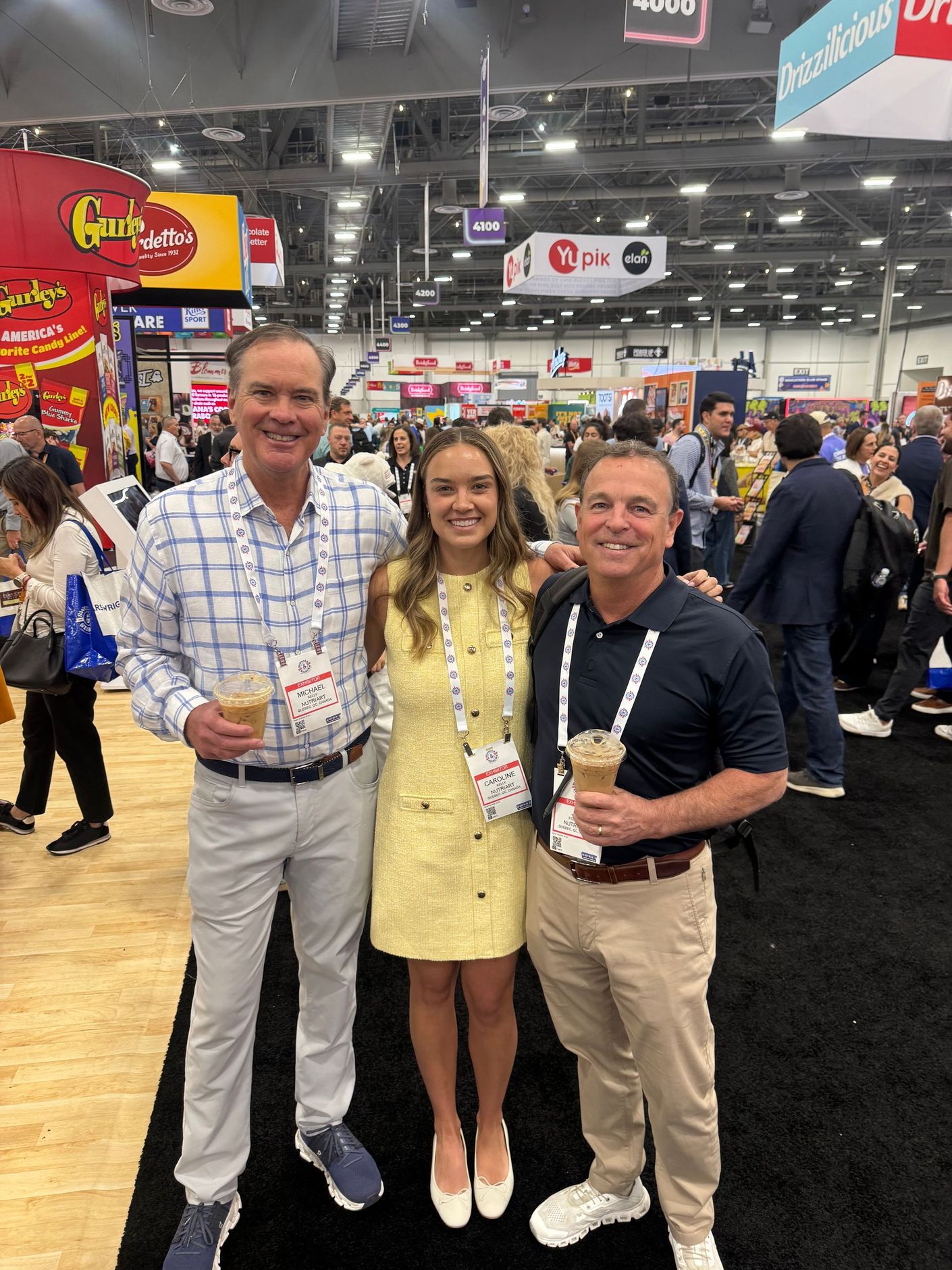 Three people smiling at an indoor trade show, holding drinks and wearing lanyards