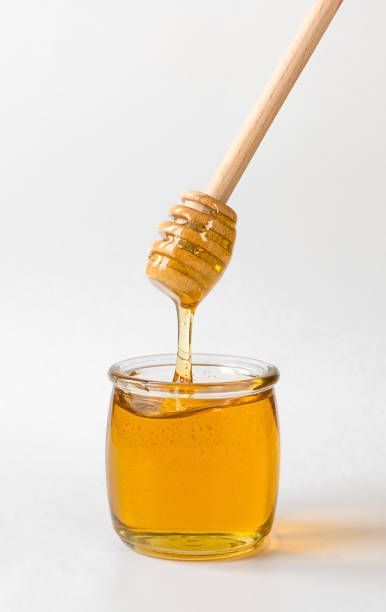 Honey is dripping from a wooden dipper into a glass jar
