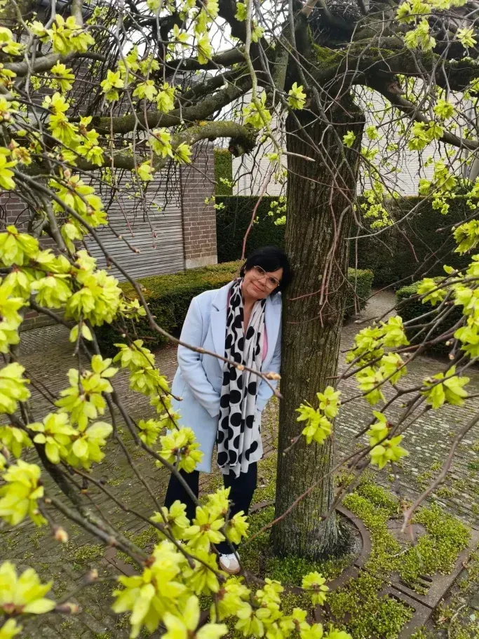 Een persoon in een witte jas staat bij een boom met heldere lentebladeren in een tuin.