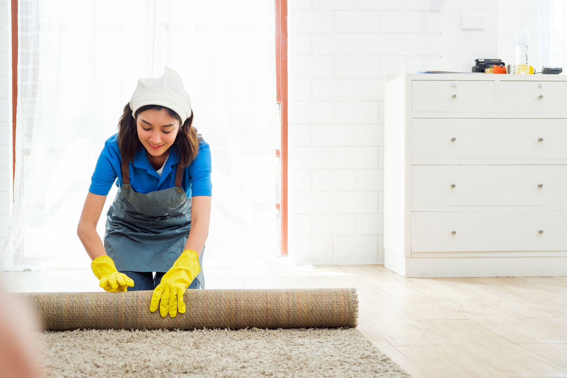 A technician in a blue uniform and yellow gloves rolling up carpet in a bright, clean living room