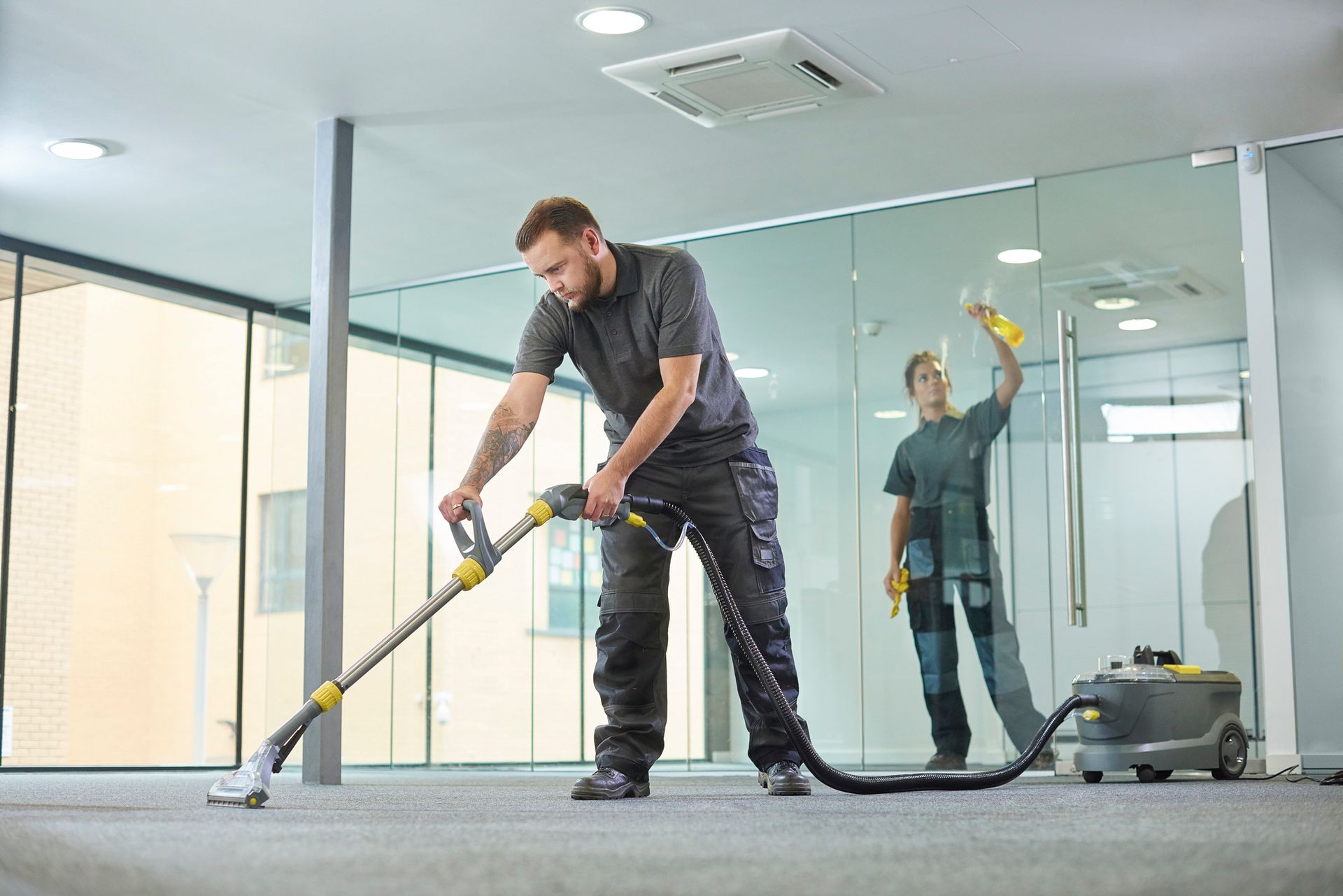 A male contractor providing carpet cleaning services, with female cleaning glass behind. A male contractor providing carpet cleaning services, with female cleaning glass behind.