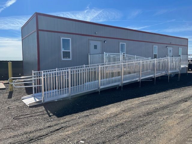 Portable gray building with white ramp and railing on a gravel lot under a clear blue sky