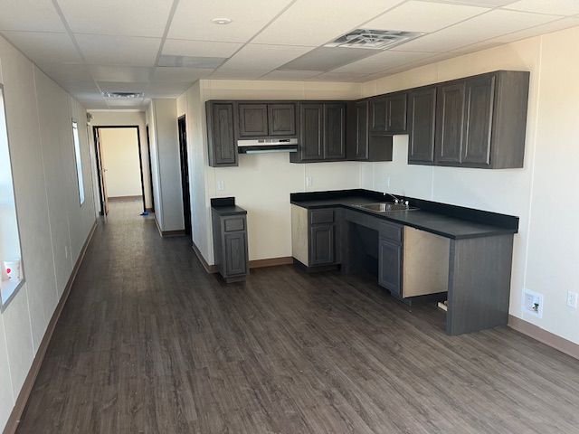 Empty office kitchenette with dark cabinets, black counters, and wood-look flooring in a long hallway.