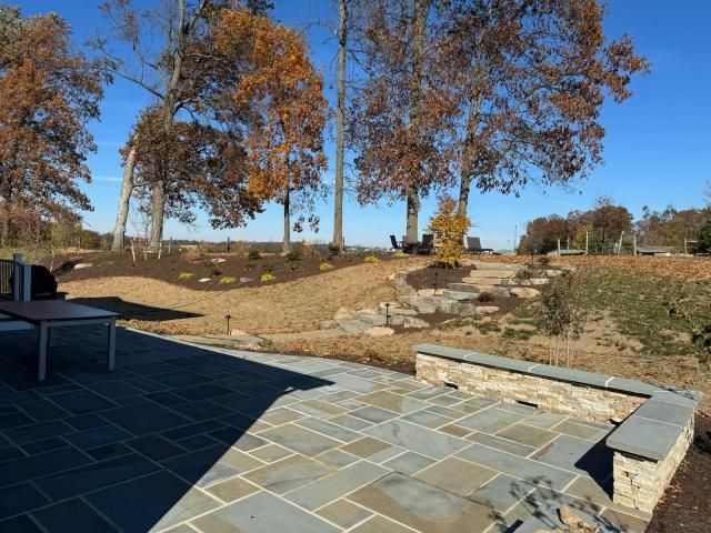 A stone fire pit sits in the center of a circular flagstone patio, connected by a path to a brick house in the background.