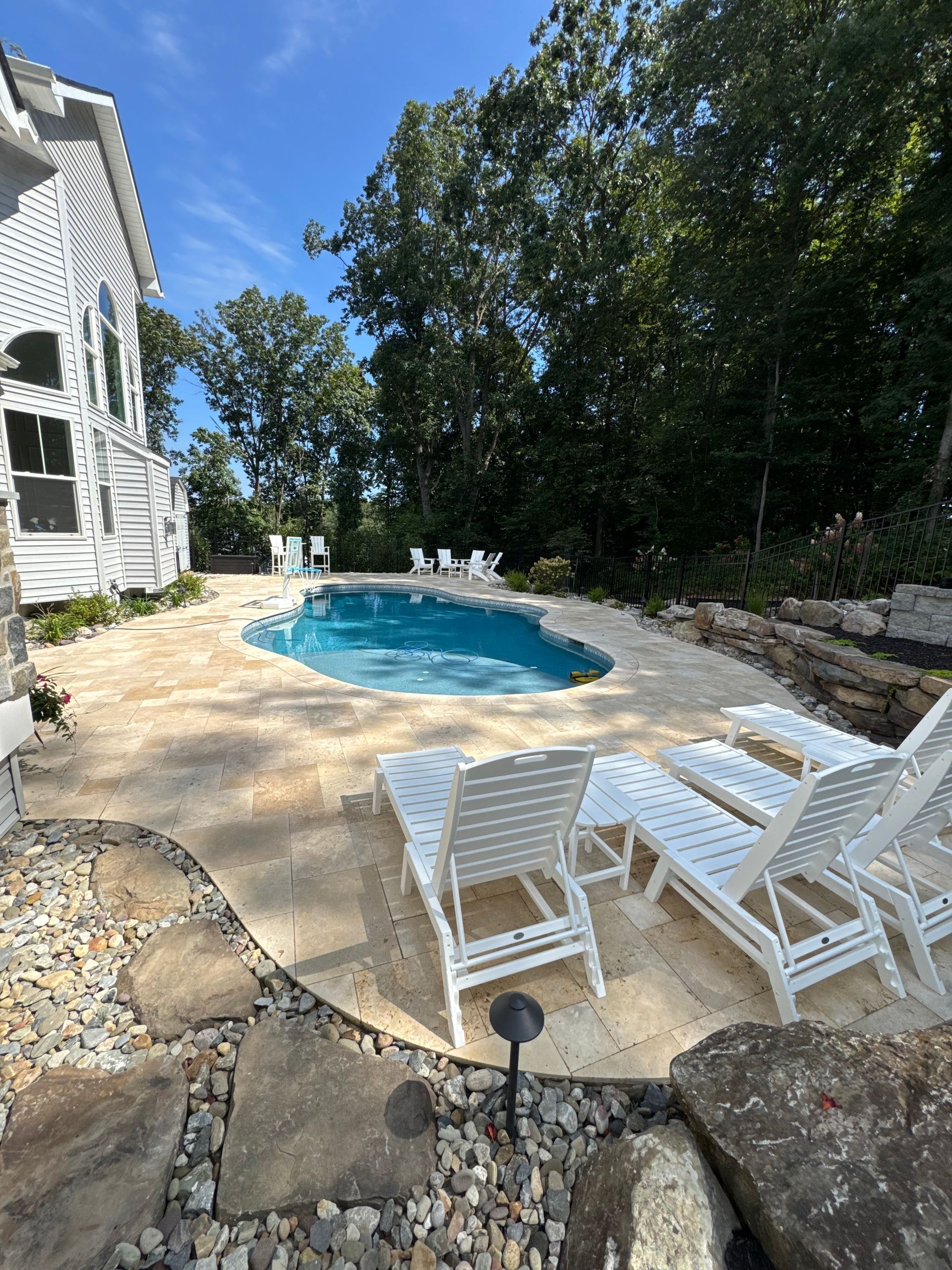 A backyard swimming pool with light stone decking, surrounded by white lounge chairs and lush trees under a sunny sky.