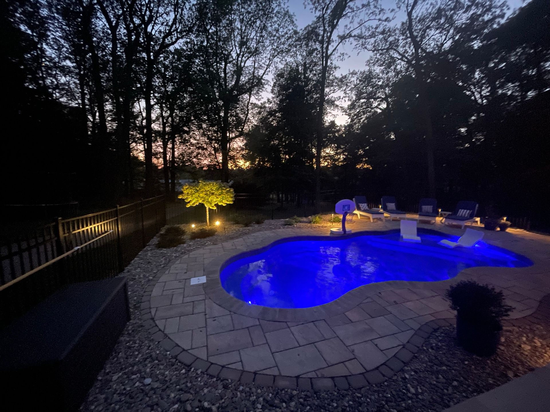 A backyard pool glows blue at dusk, surrounded by a stone patio, landscape lighting, and trees against a sunset sky.