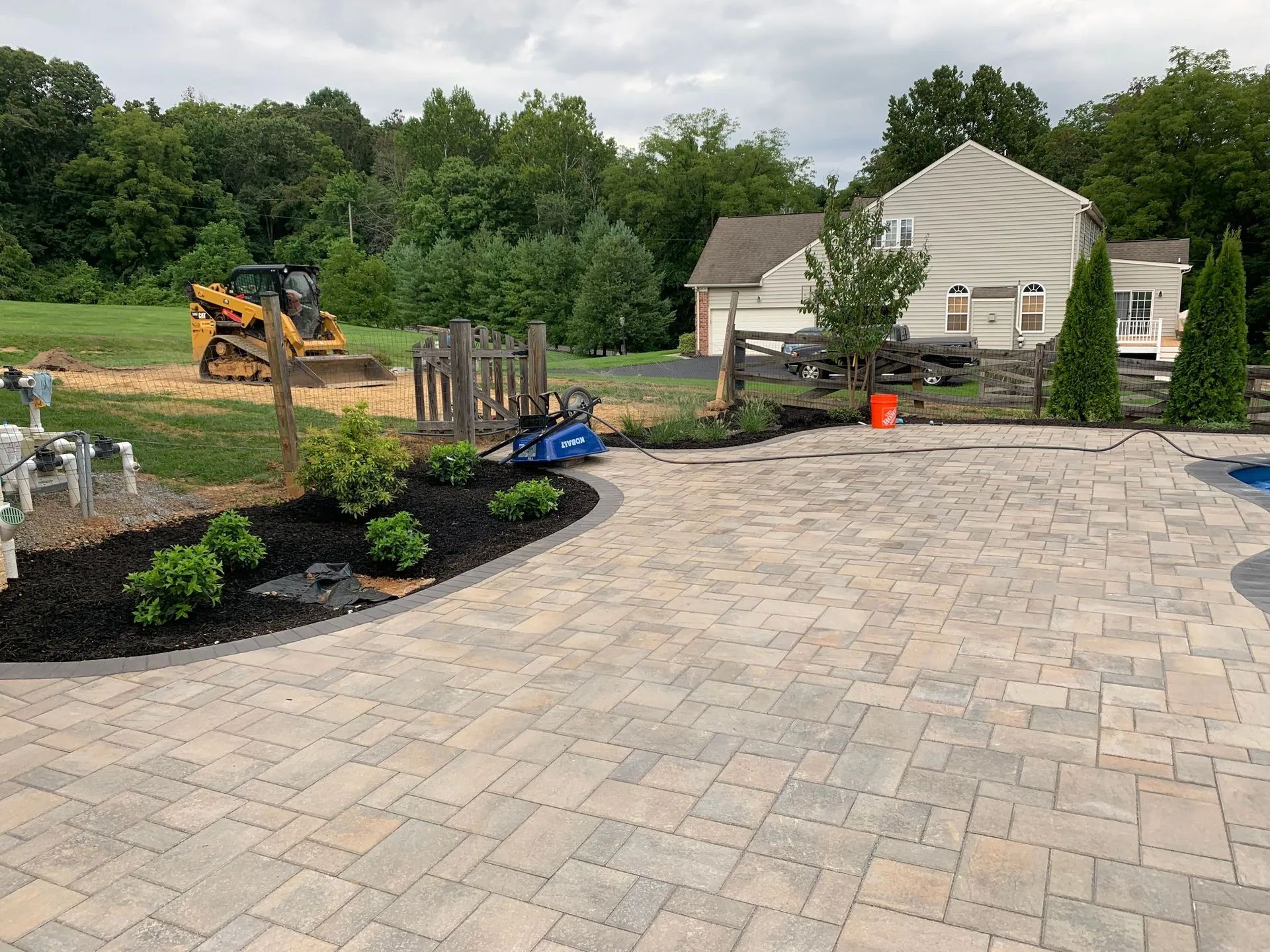 Newly paved patio with garden beds, a small excavator, and a house in the background