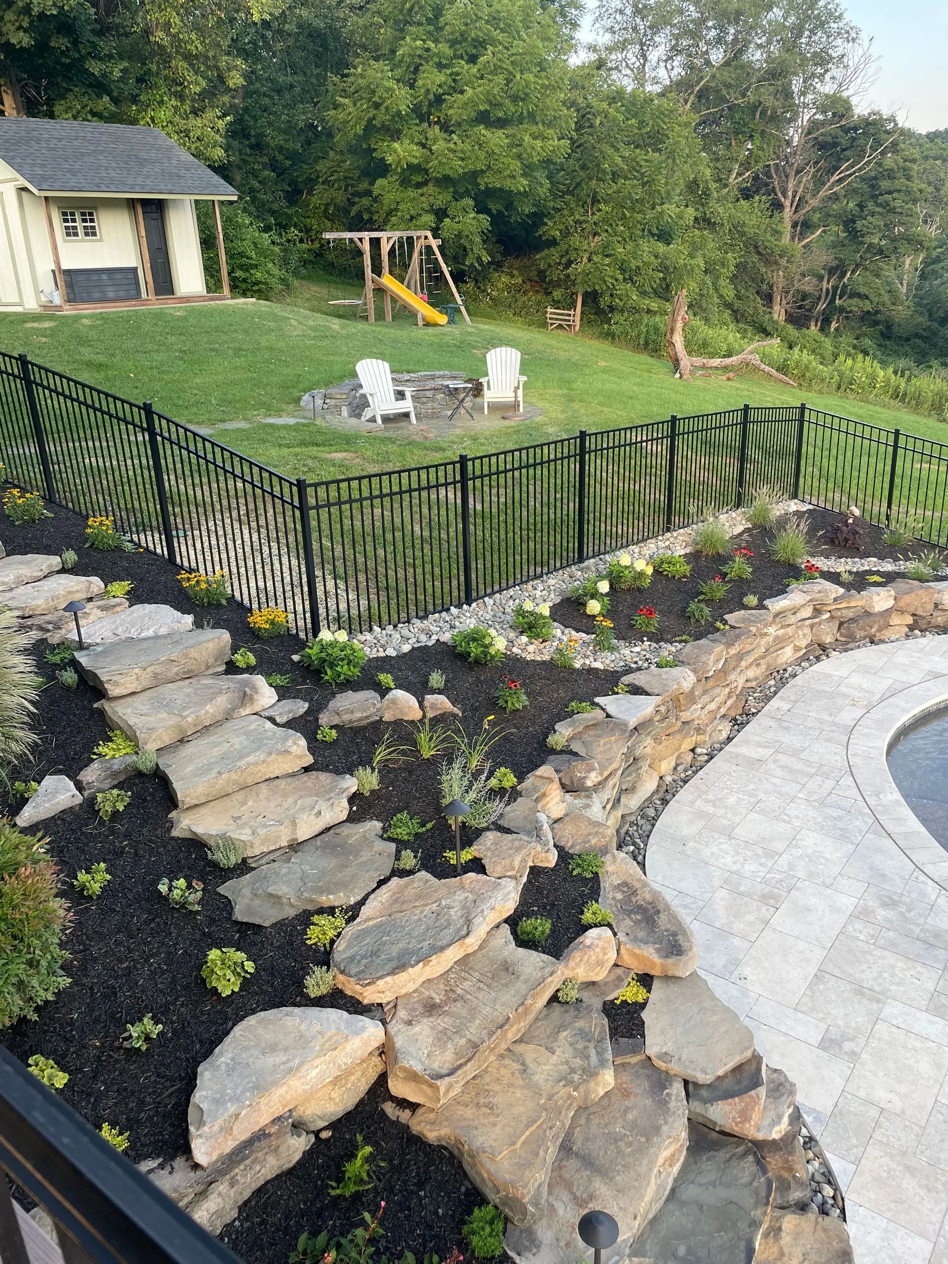 Backyard landscape with stone steps, black fence, lawn, flower beds, patio, and a swing set at the top.