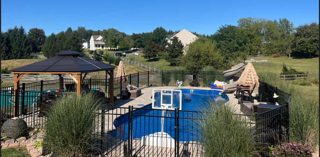 A backyard swimming pool with a gazebo, slide, and basketball hoop, enclosed by a black metal fence on a sunny day.