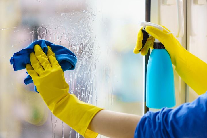 Gloved hands cleaning a glass window with blue cloth and spray bottle