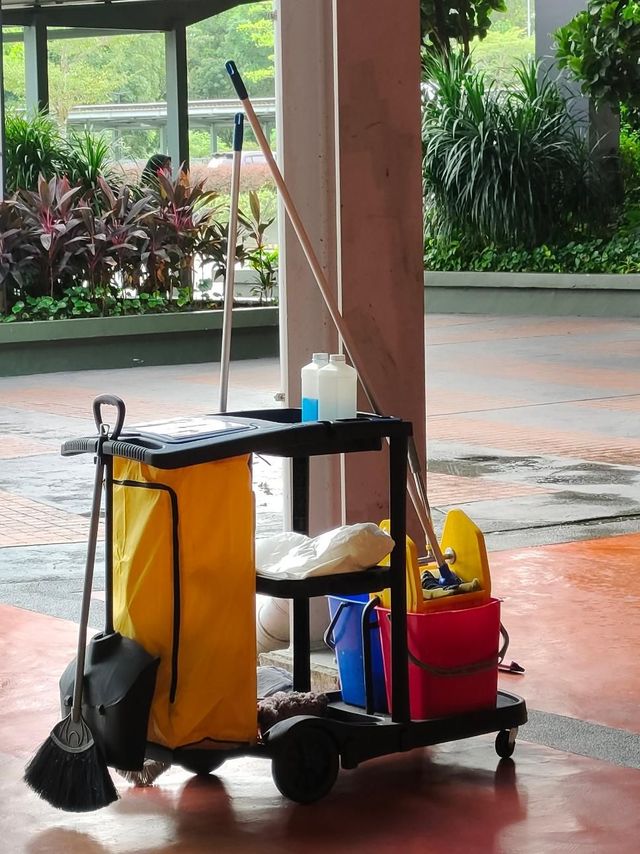 Janitorial cart with cleaning supplies, mop, and buckets on a wet tiled floor under a covered walkway