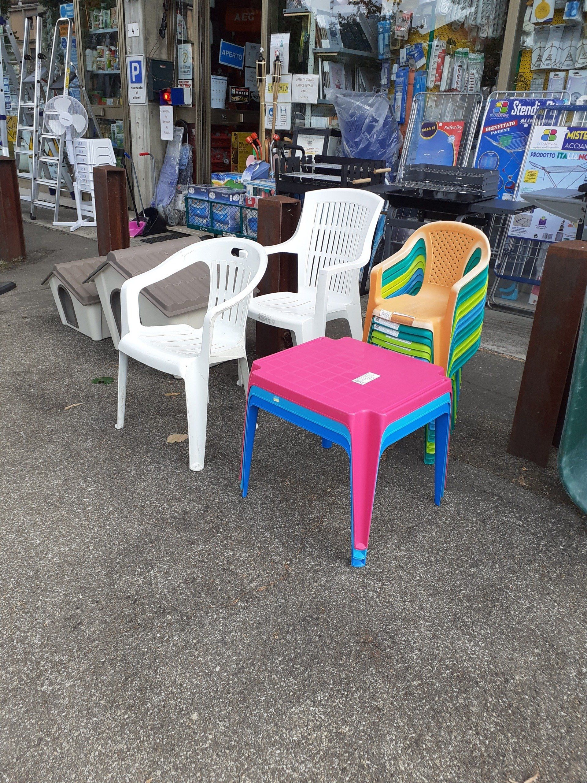 Quattro sedie di plastica e uno sgabello rosa all'esterno di un'esposizione di negozio, di fronte a una vetrina ingombro di oggetti.