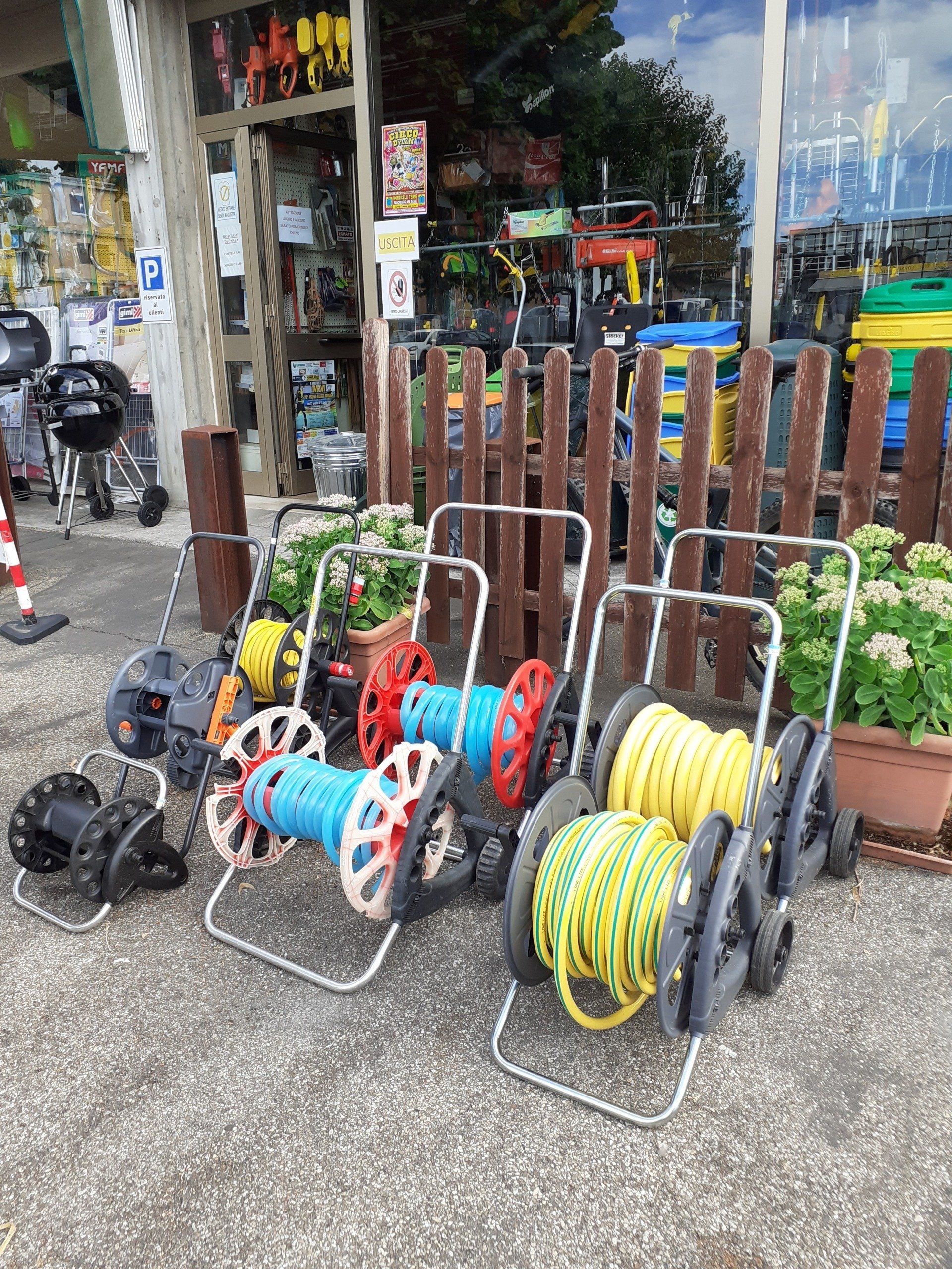 Tubi da giardino colorati arrotolati su carrelli davanti a un negozio, con una recinzione di legno sullo sfondo.