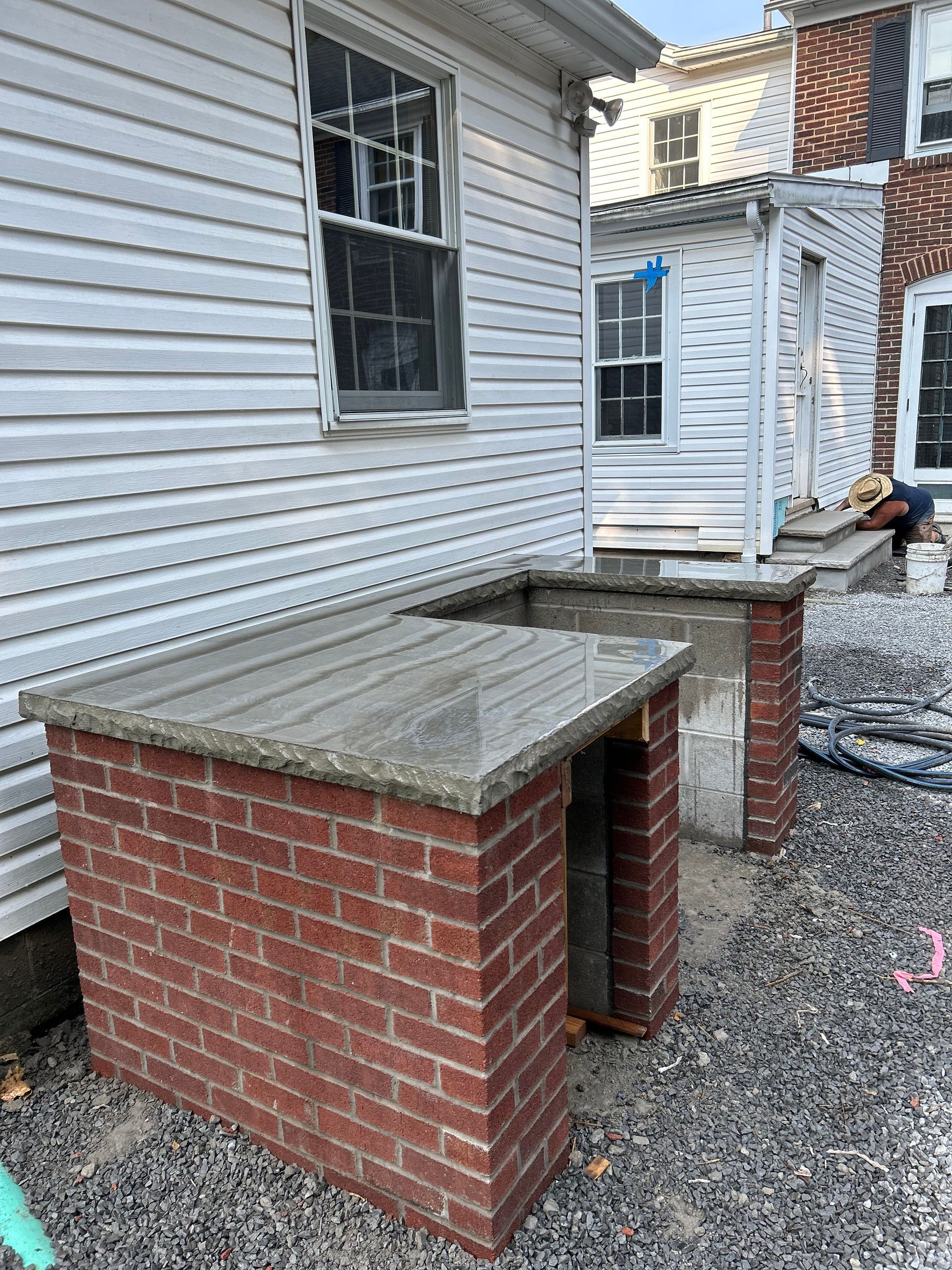 Two unfinished brick outdoor kitchen counters with stone tops stand on a gravel ground next to a white siding house.