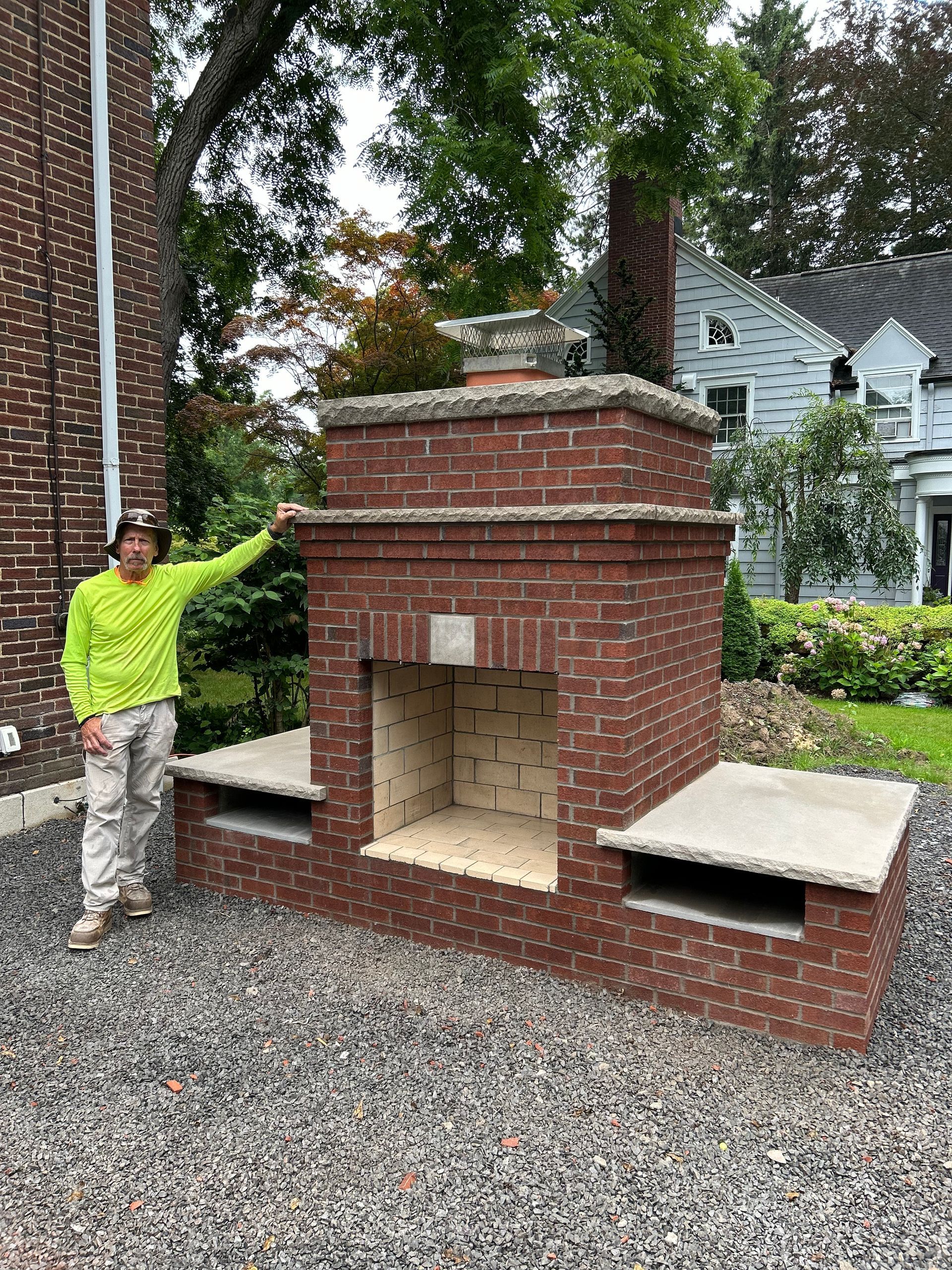 A person in a neon yellow long-sleeved shirt standing next to a newly built red brick outdoor fireplace with side benches.
