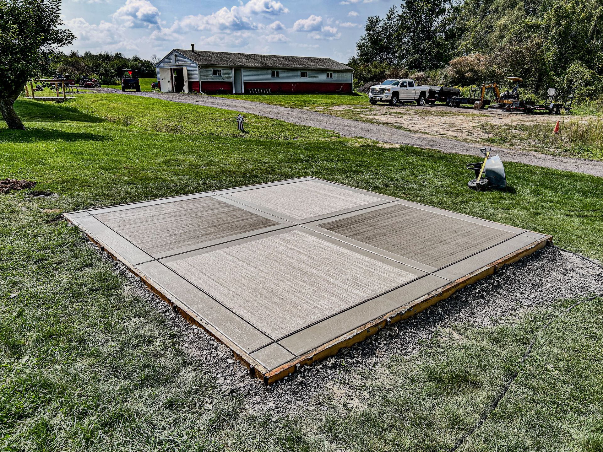 A square, flat-topped gravel foundation with a wooden frame sits in a grassy yard, with a white barn in the distance.