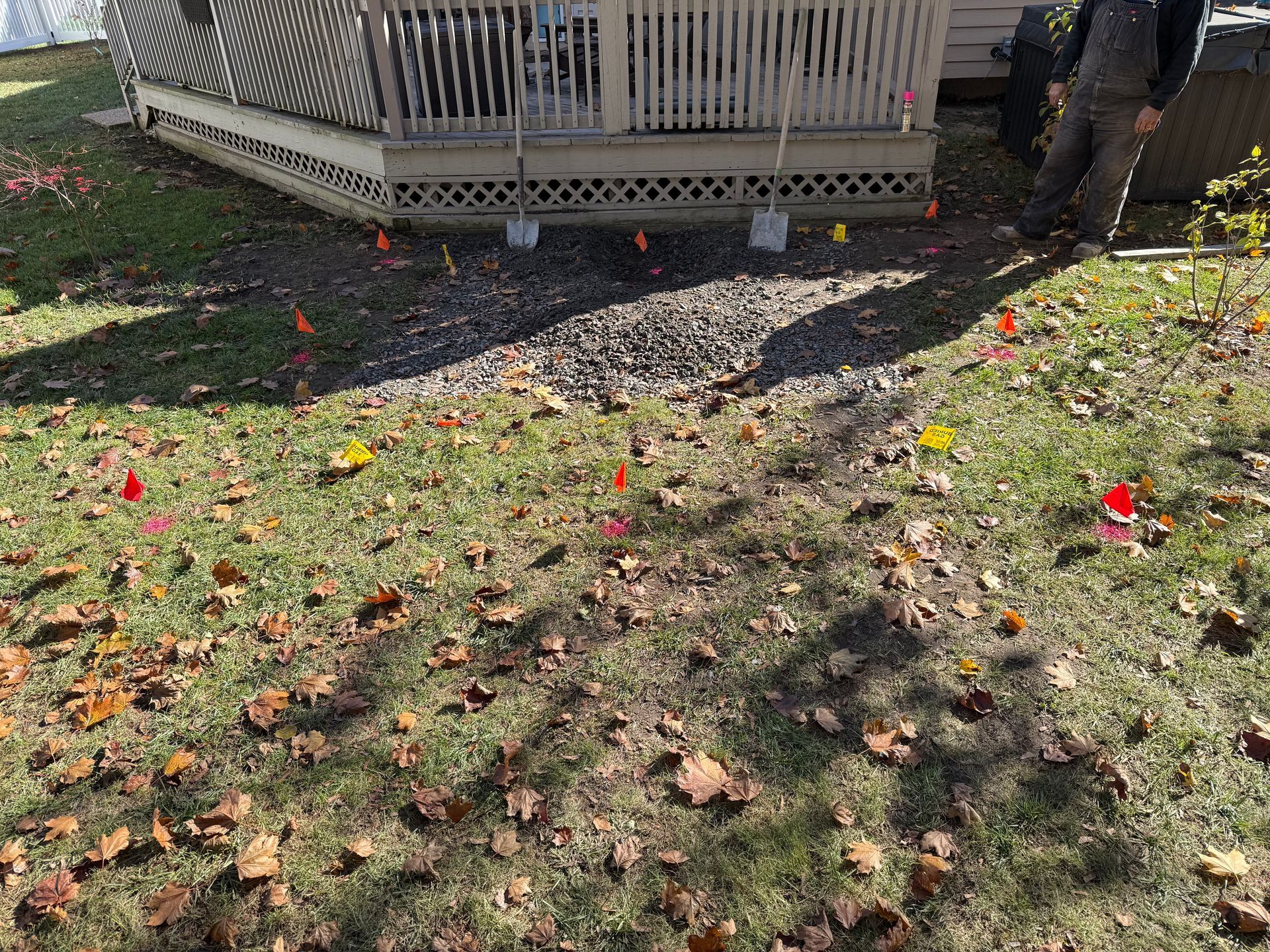 A person stands near a deck with a gravel area marked by small flags in a yard covered with autumn leaves.
