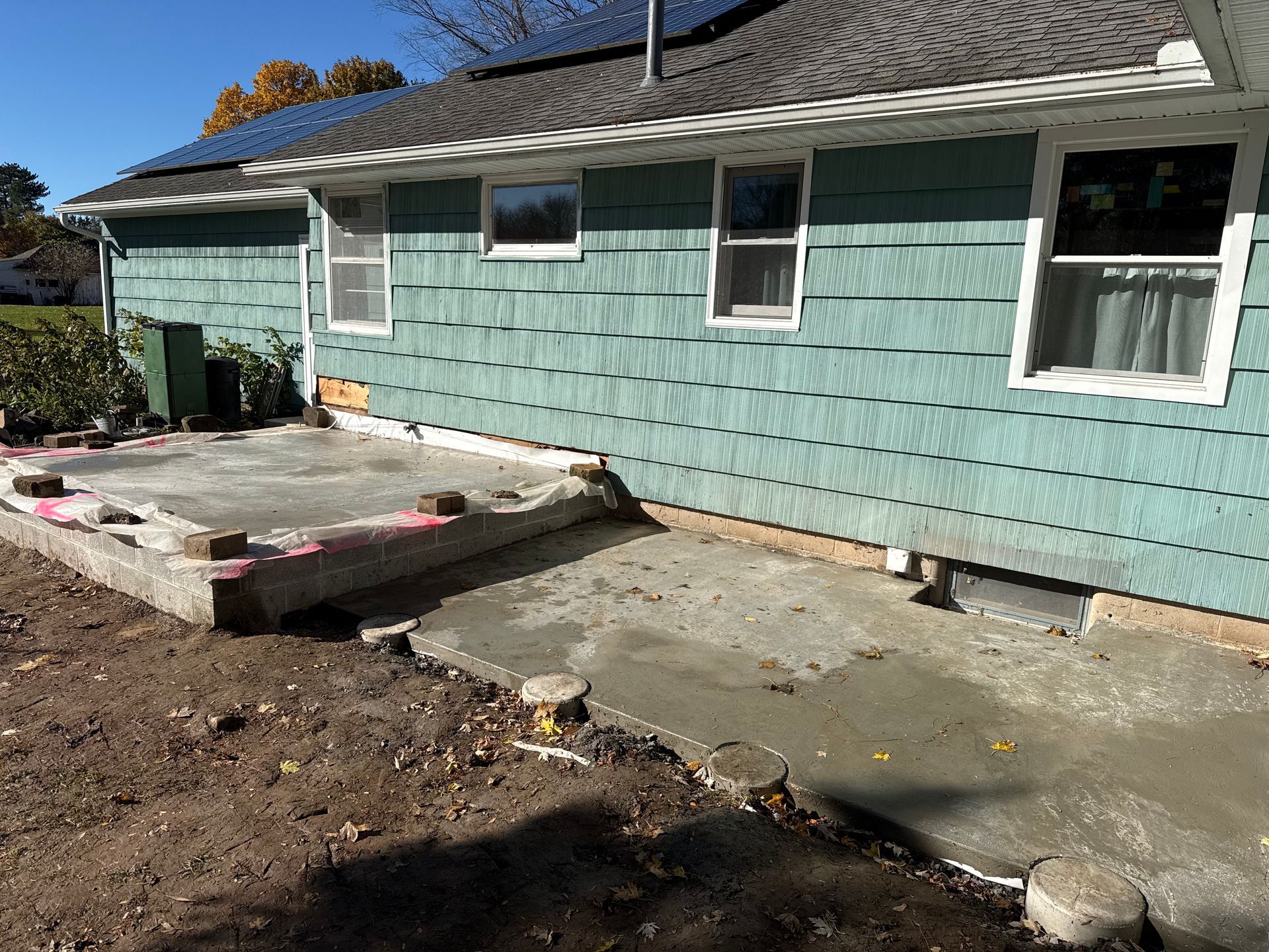 A freshly poured concrete patio slab outside the light blue shingled wall of a house, with several concrete pier footings.