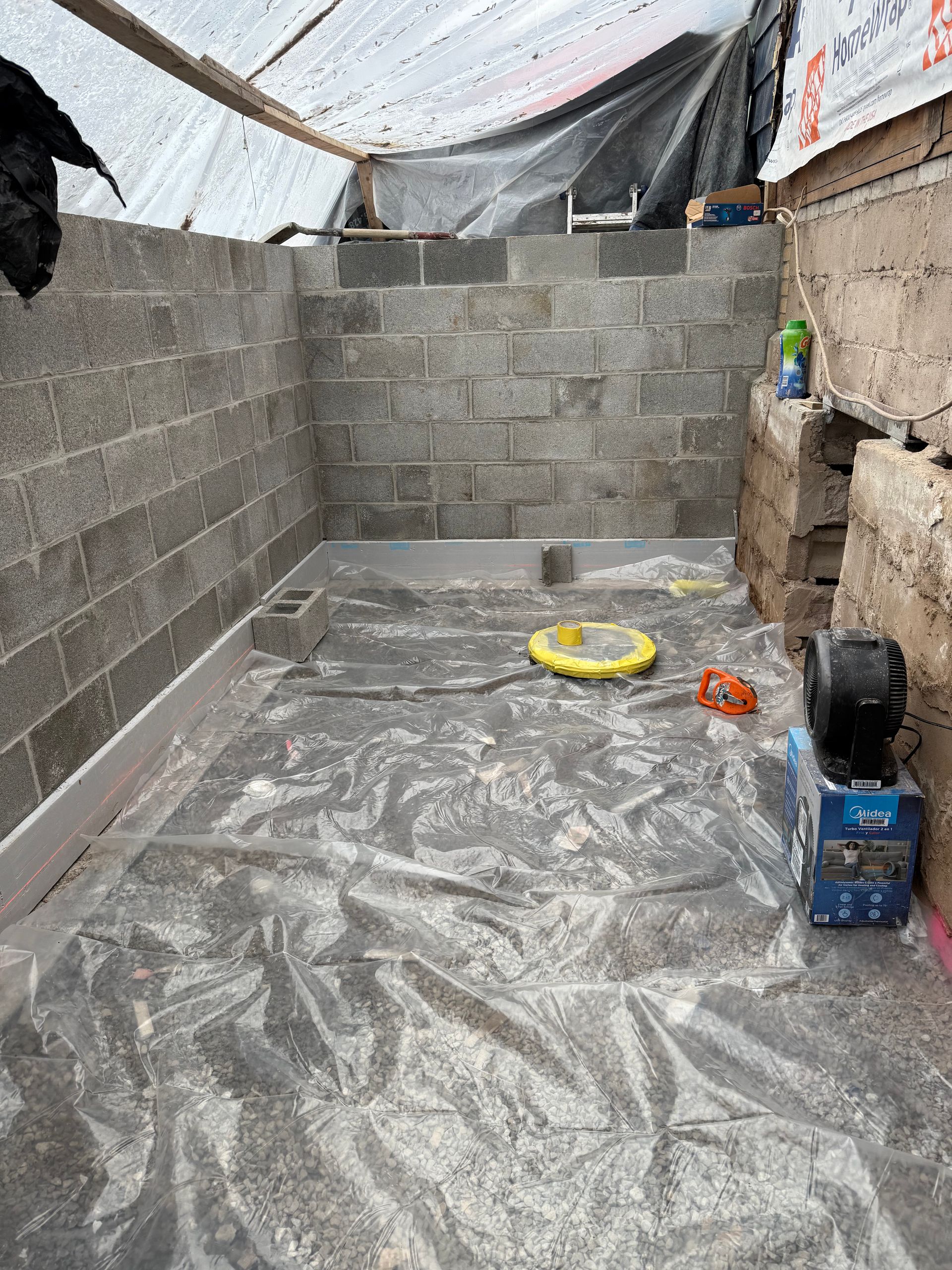 An unfinished room with cinder block walls, a plastic-covered floor, and construction equipment under a tarp roof.