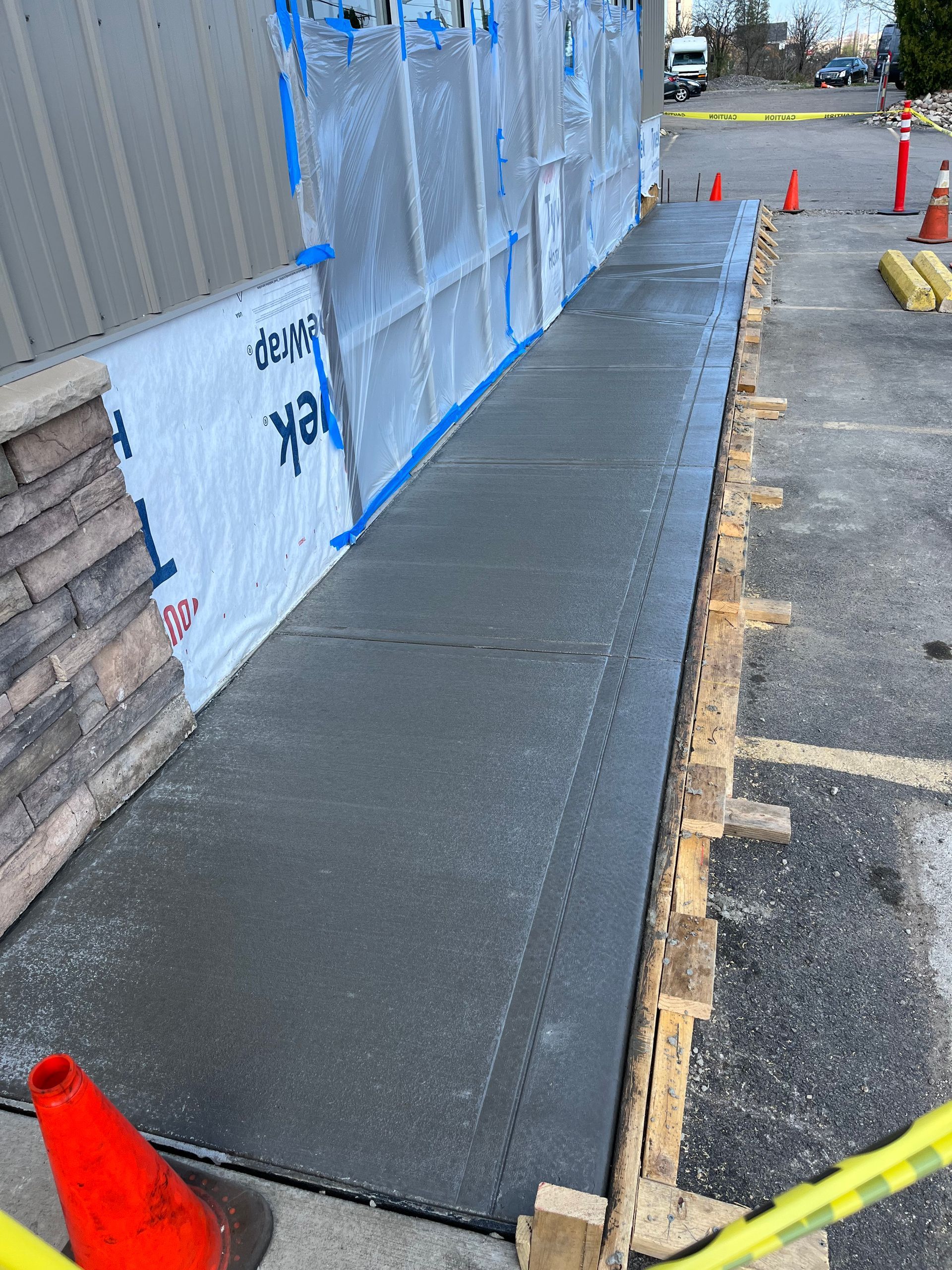 A freshly poured concrete sidewalk runs alongside a building under construction, bordered by wooden forms and orange cones.