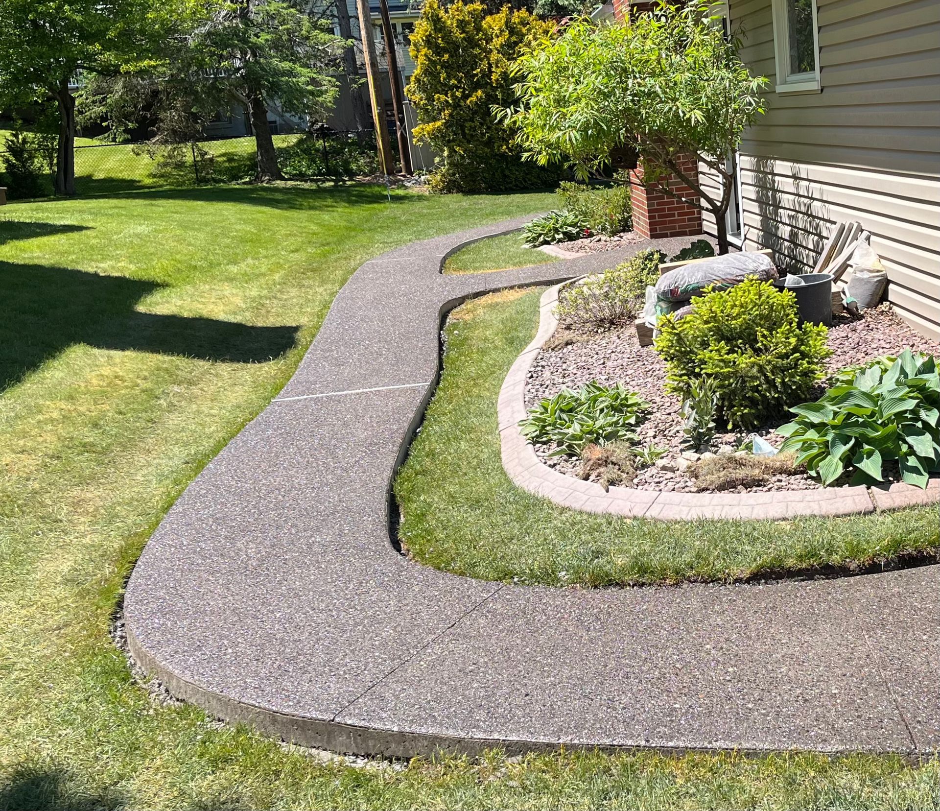 A textured concrete walkway curves through a lawn beside a residential house with a landscaped garden bed.