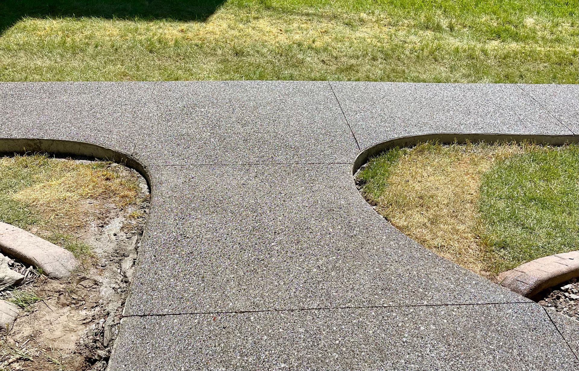 A textured concrete walkway forms a T-junction between two grassy lawn areas in a sunlit outdoor space.