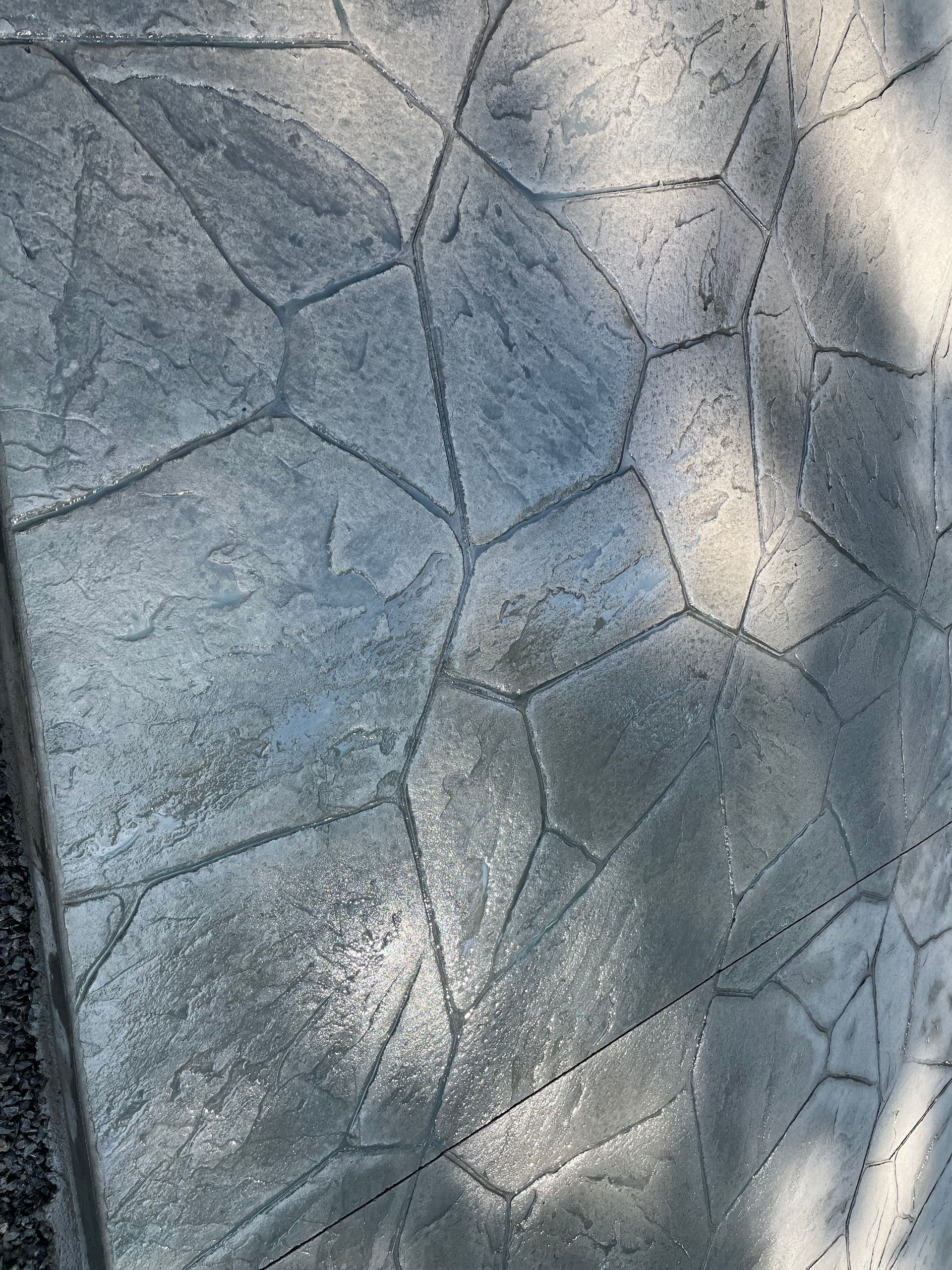 Gray concrete stamped with a flagstone pattern, featuring dark grout lines and a textured surface under dappled sunlight.
