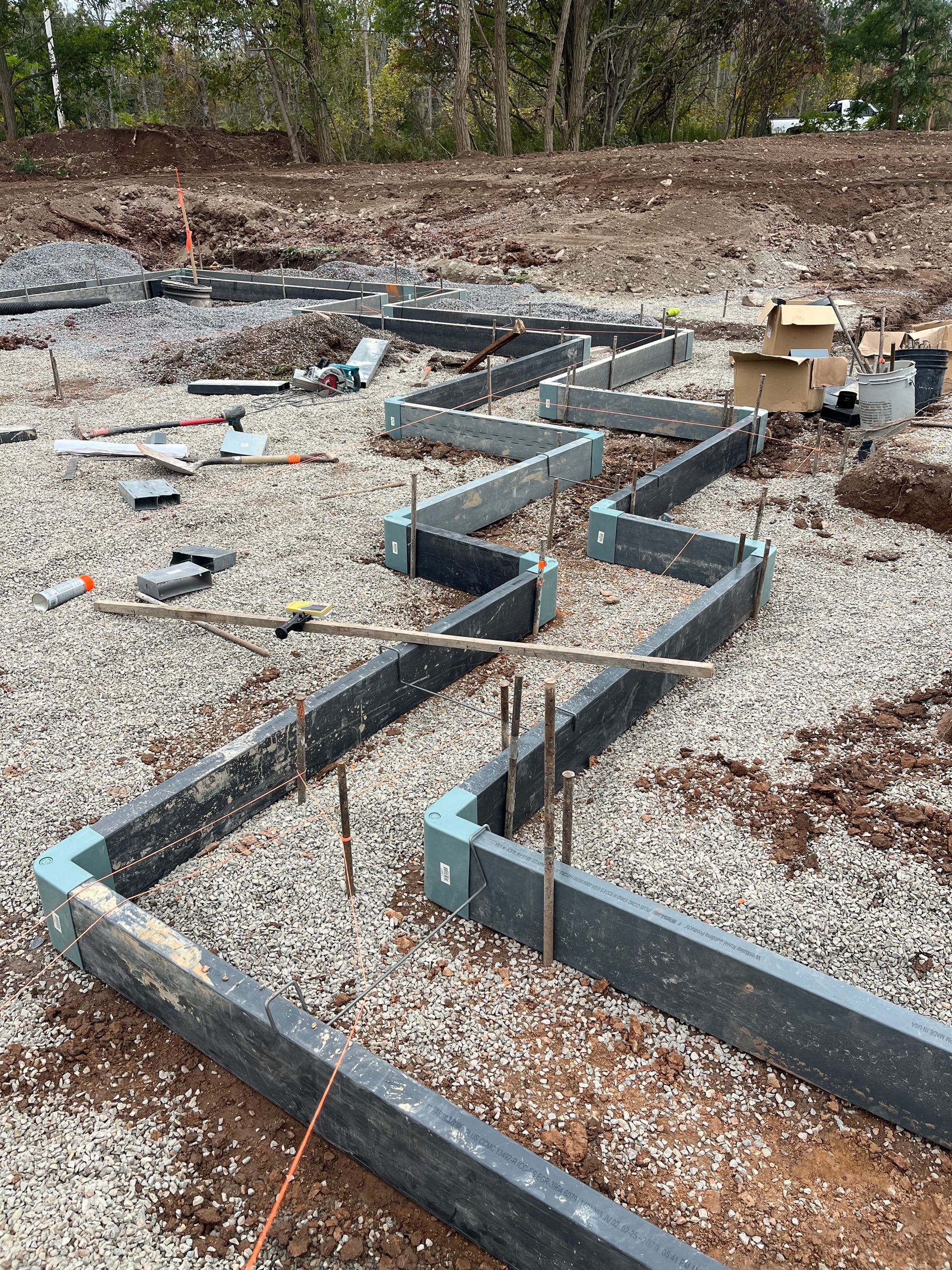 Zigzagged dark gray foam forms set on a gravel bed for a concrete foundation, with rebar stakes and tools nearby.