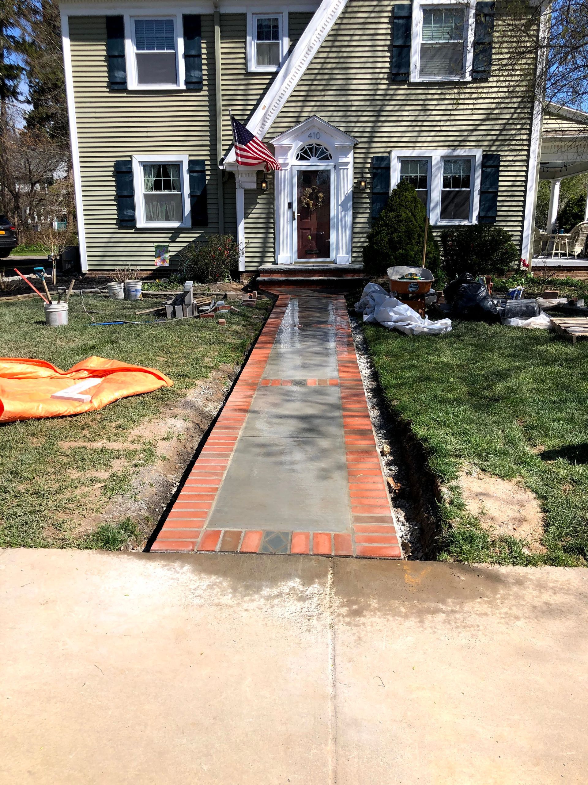 A newly installed concrete walkway with a red brick border leads to the front entrance of a two-story beige house.