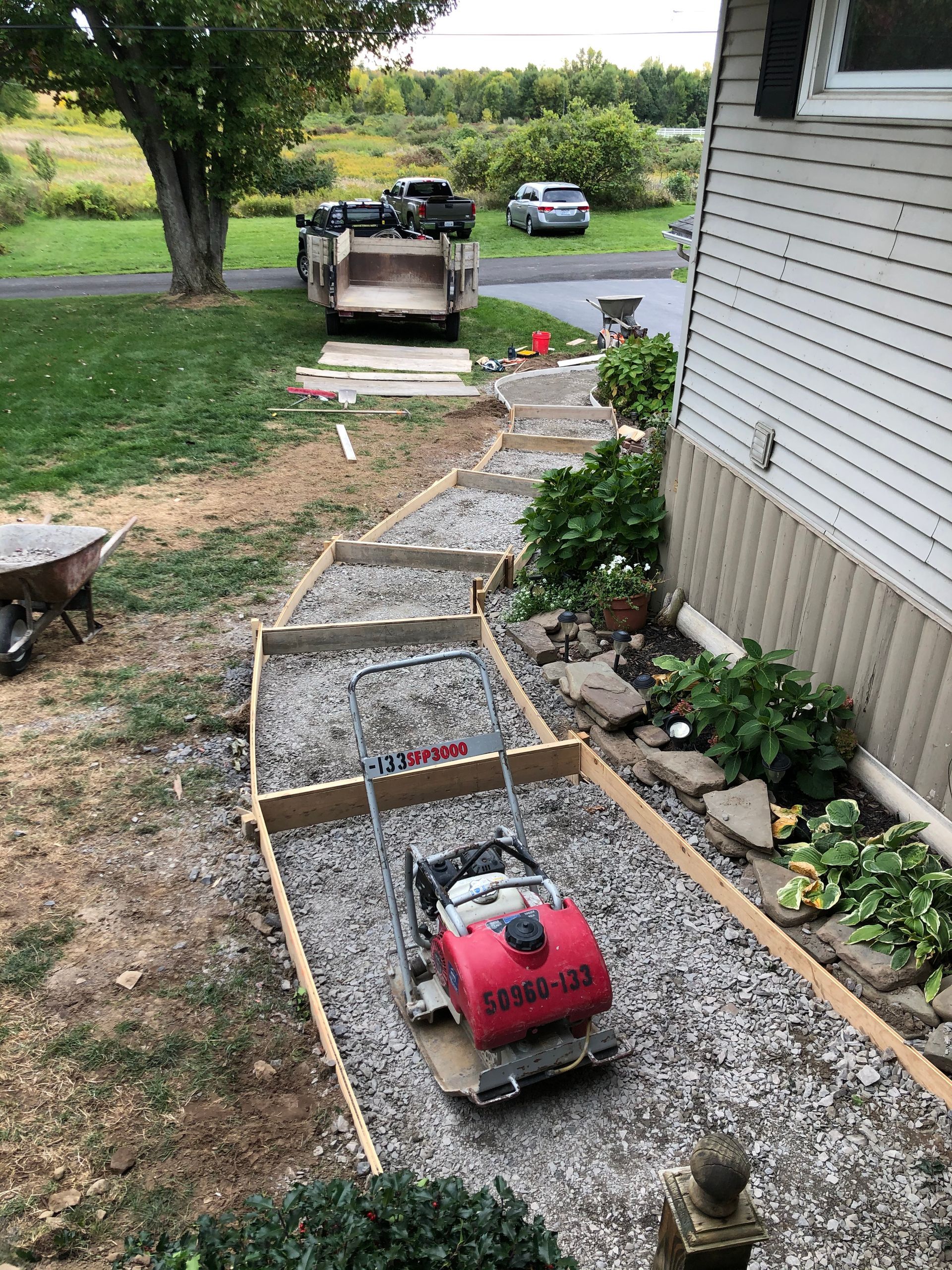 A winding gravel path under construction with wooden forms and a red plate compactor, alongside a house with siding.