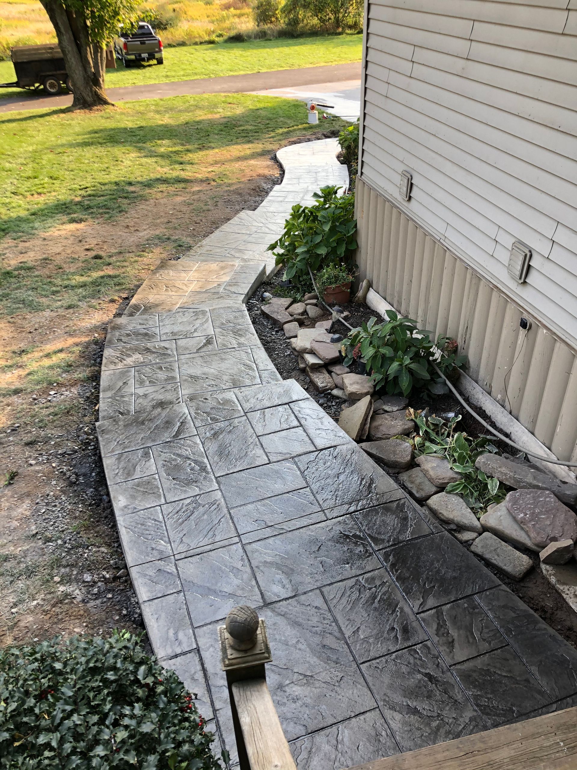 A stamped concrete walkway with a tile pattern curves along the side of a house, leading toward a grassy yard.