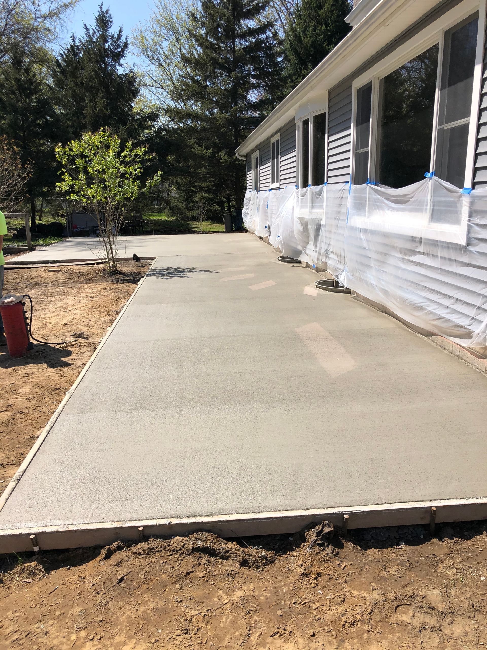 A newly poured concrete patio runs along the side of a house wrapped in protective plastic.