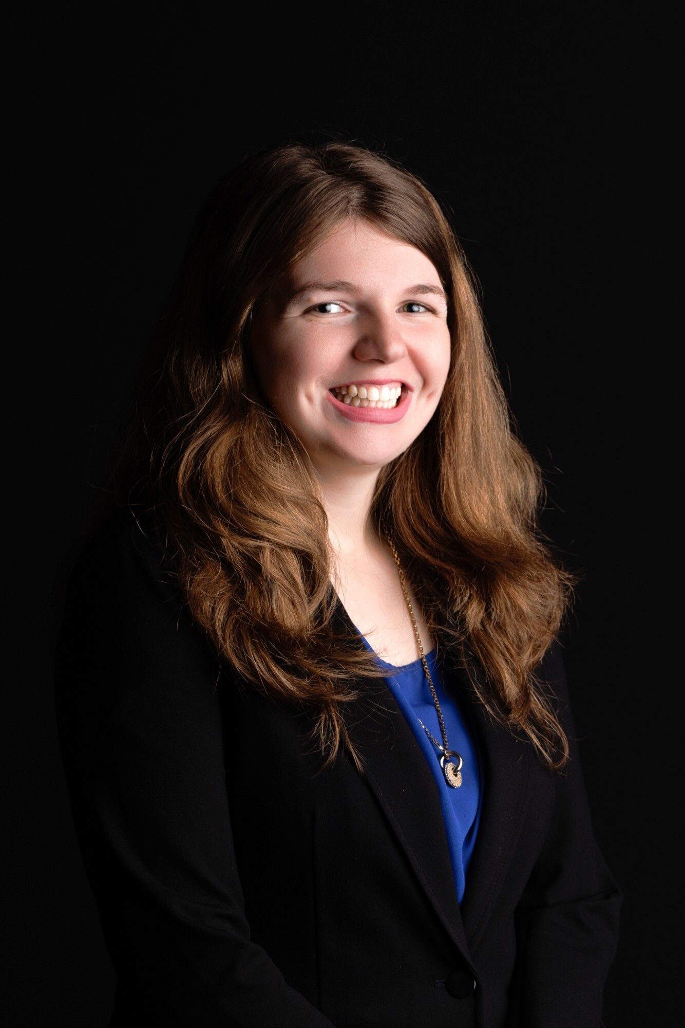 Young woman smiling, wearing a black blazer over a blue top, with long brown hair, against a black background.