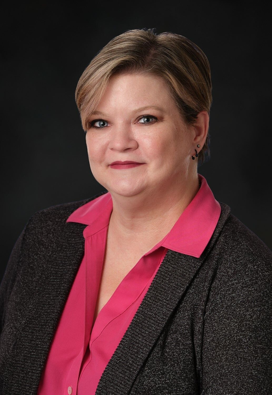 Portrait of a smiling person with black glasses and dark hair, wearing a burgundy blazer on a black background