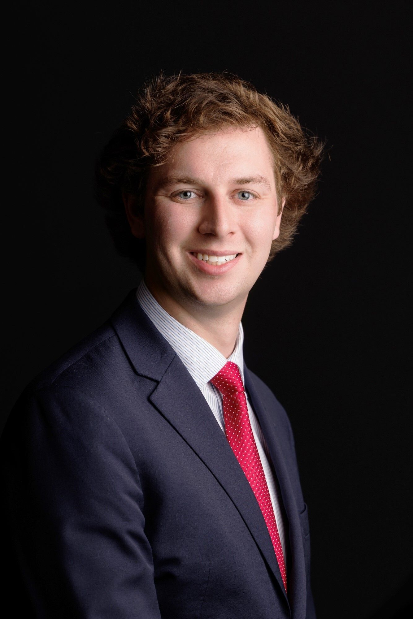 Man in navy suit, red tie, smiling against a black background.