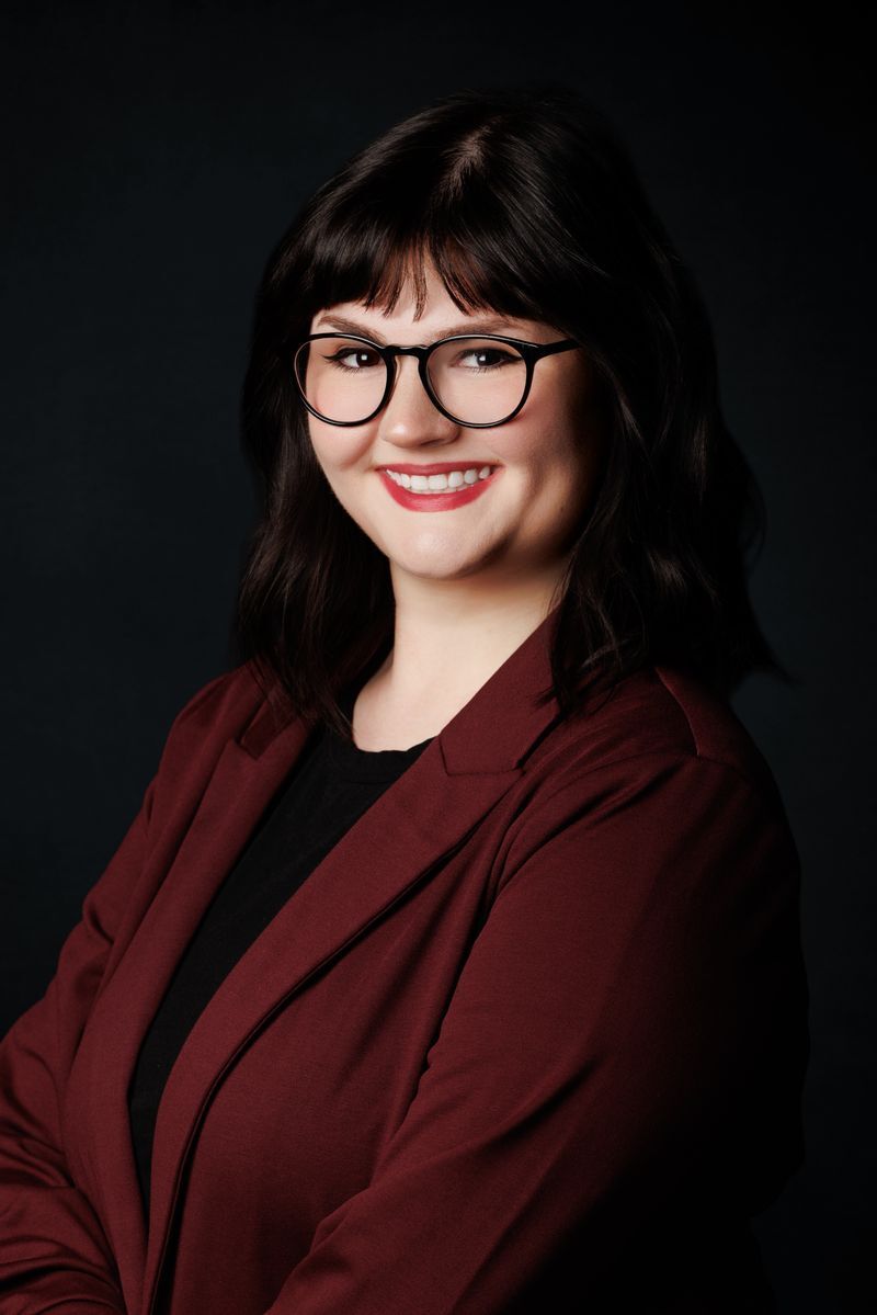 Portrait of a smiling person with black glasses and dark hair, wearing a burgundy blazer on a black background