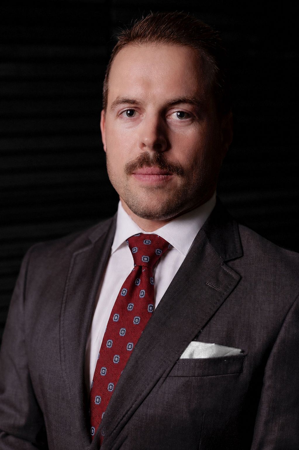 Studio portrait of a man in a dark suit, red patterned tie, and white pocket square against a black background
