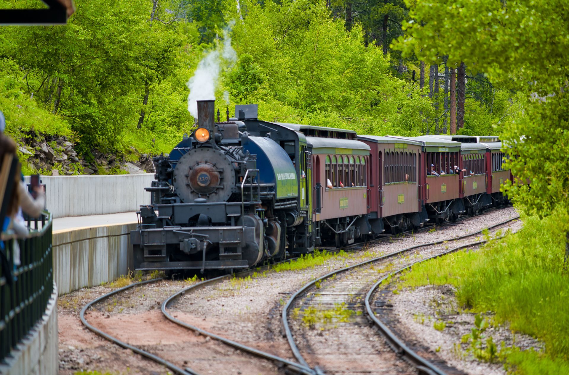 Steam locomotive pulling passenger cars around a curved track through a green wooded area