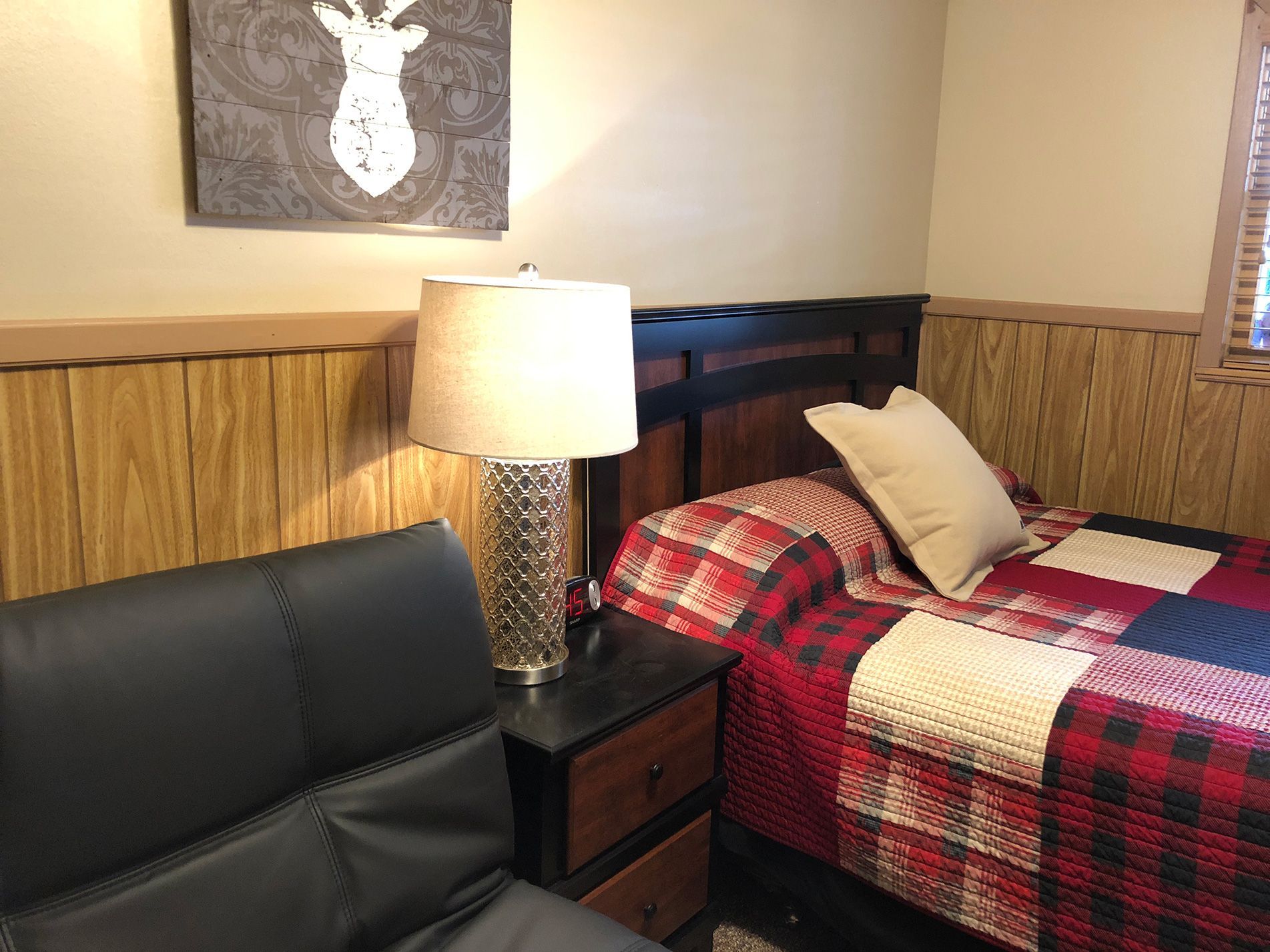 Bedroom with a plaid quilt, lamp, nightstand, and black sofa.  Wall art depicts a deer.