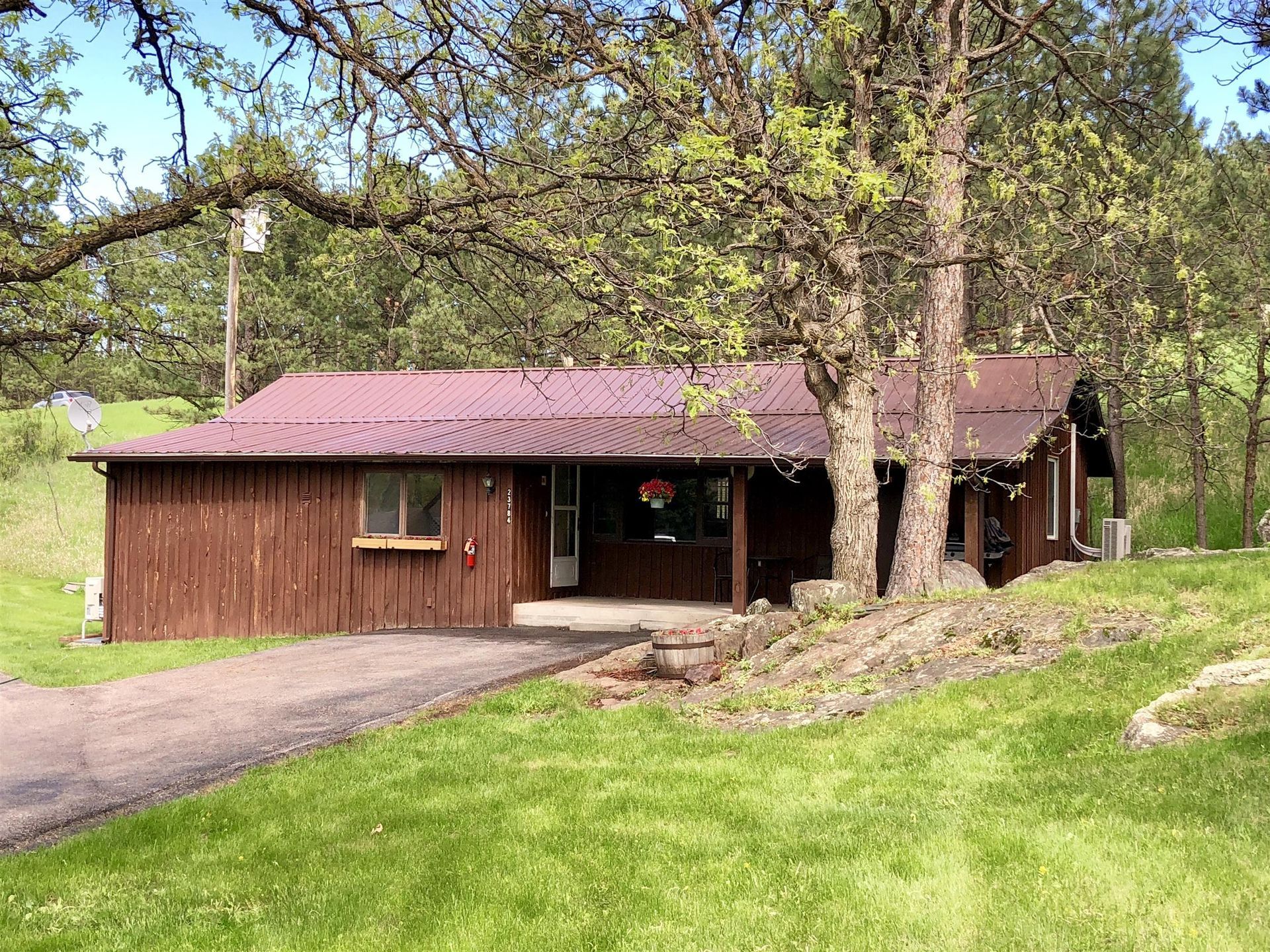 Brown cabin with dark metal roof sits on a grassy hill; a tree shades the porch.