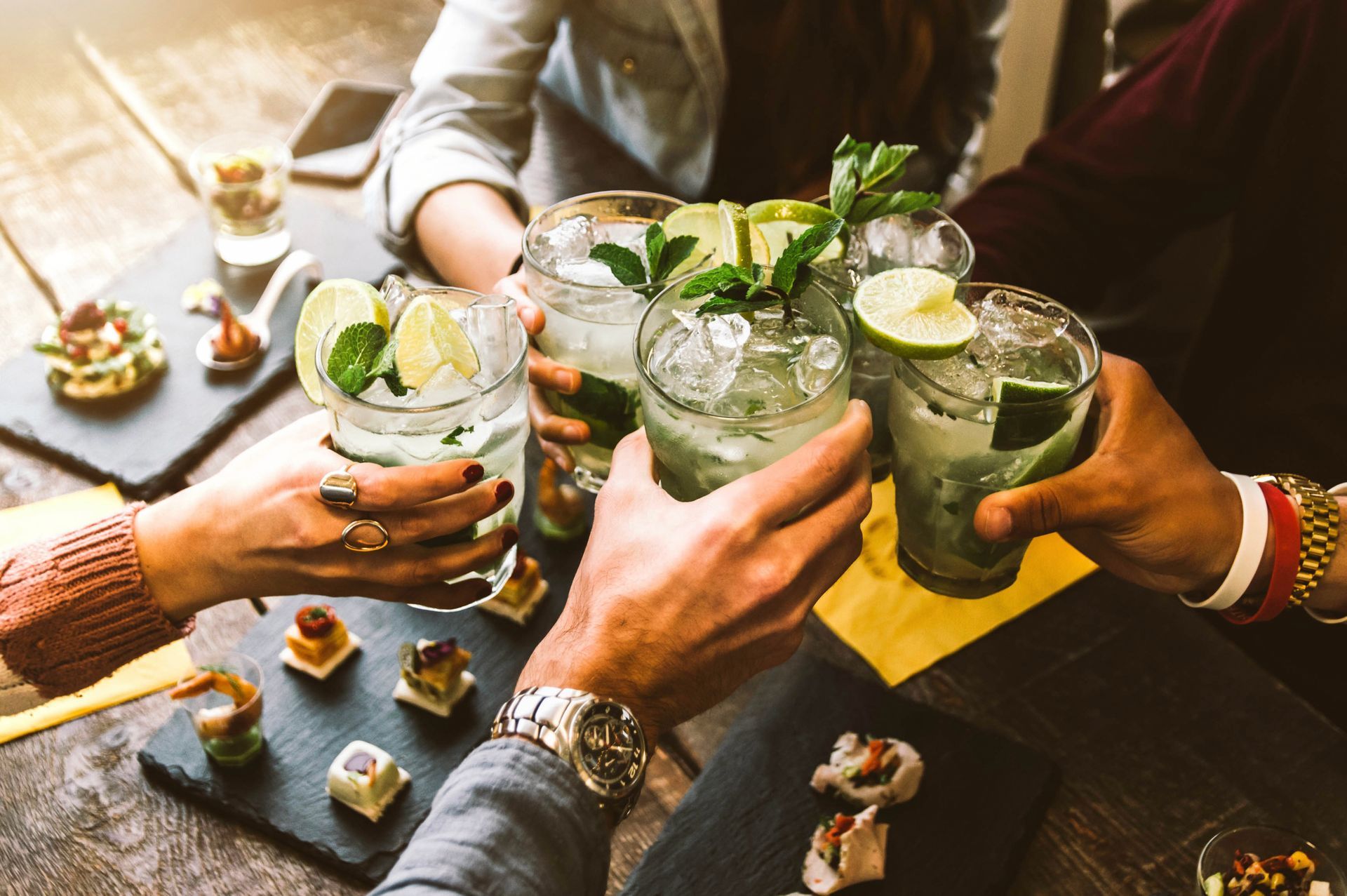 People toasting mojitos with appetizers on a dark wooden table.