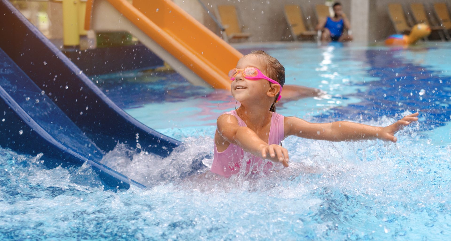 Girl wearing pink swimsuit and goggles exits a water slide into a pool, smiling with arms outstretched.